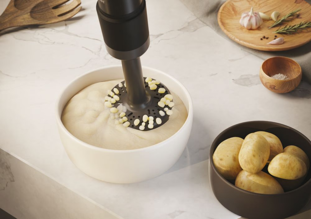 Ein Stabmixer in einer Schüssel mit Kartoffelpüree, daneben Kartoffeln und Knoblauch auf Marmortisch.