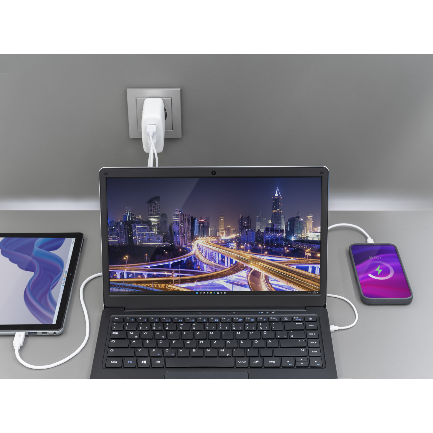 Een laptop, tablet en telefoon aangesloten op een stopcontact. De laptop toont een stadsgezicht.