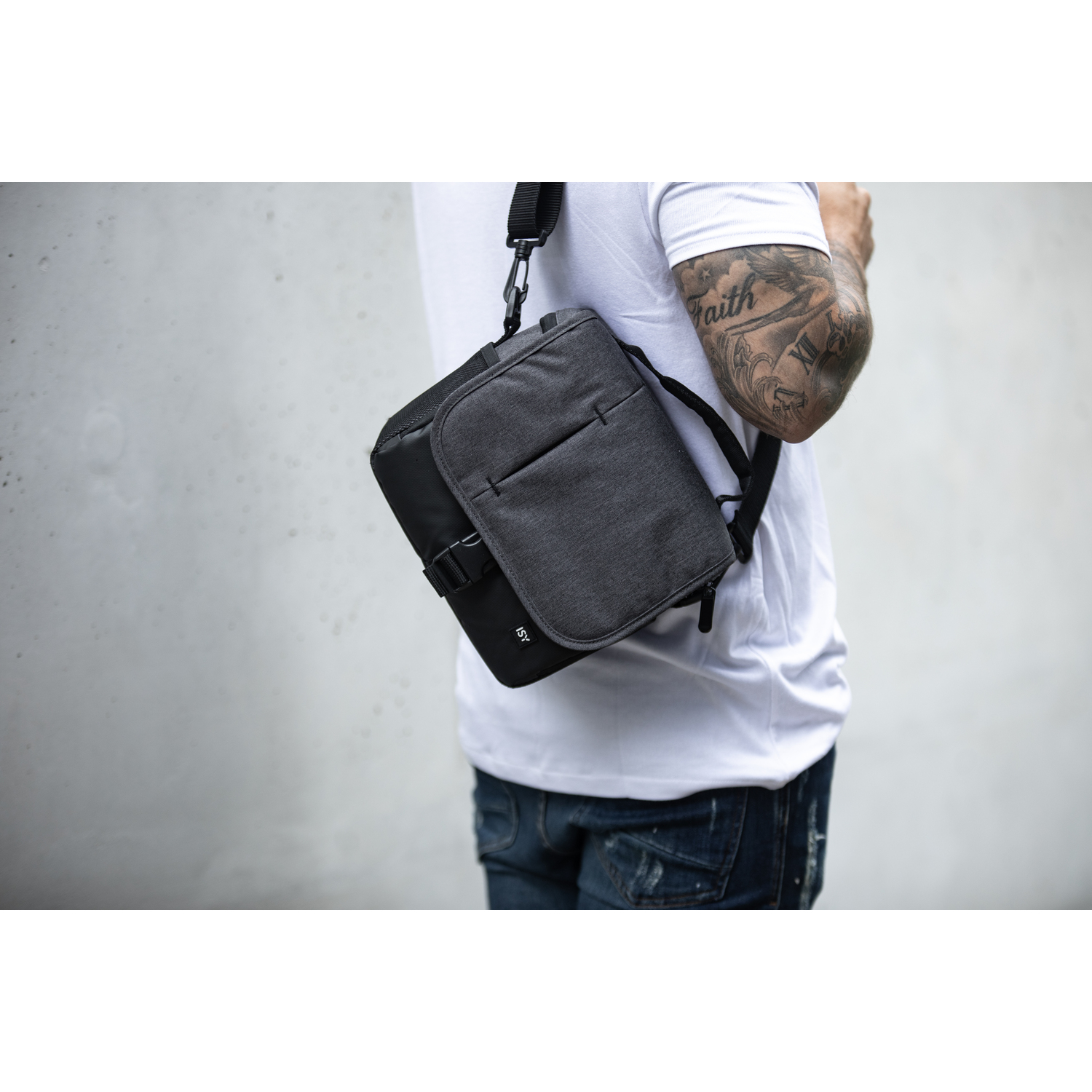 Personne portant un sac messager gris avec une sangle noire, sur un tee-shirt blanc.