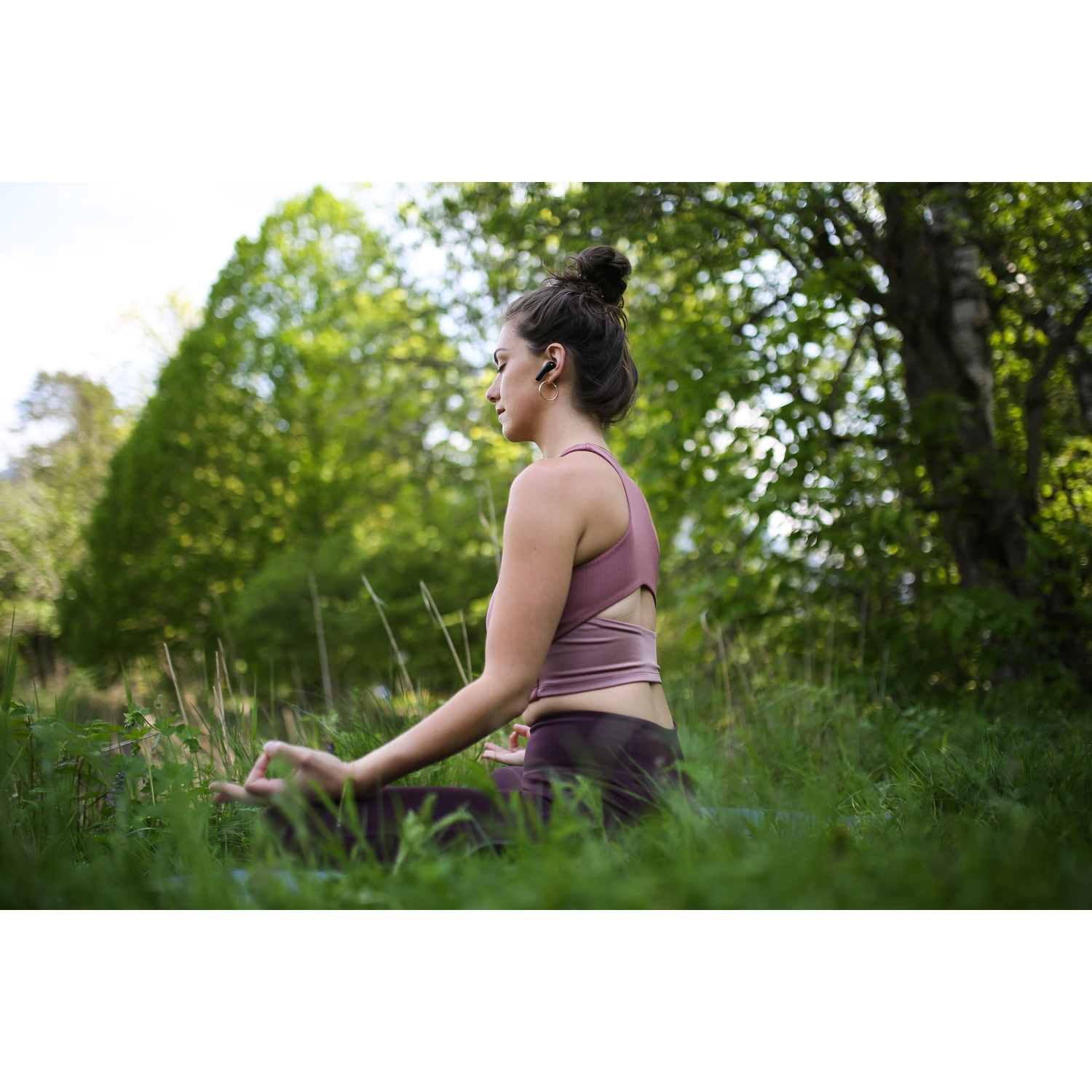 Vrouw in een park, mediterend, met oordopjes, omringd door groen gras.