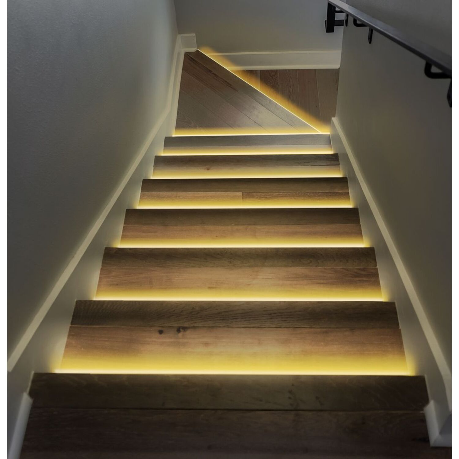 Houten trap met gele LED-verlichting, die omhoog gaat. Witte muren en een leuning zijn zichtbaar.