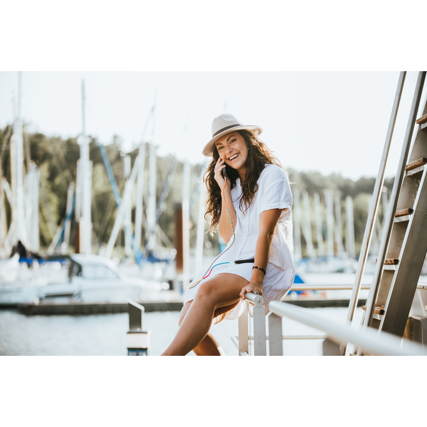 Femme en robe blanche et chapeau souriant tout en parlant au téléphone dans un port de plaisance.