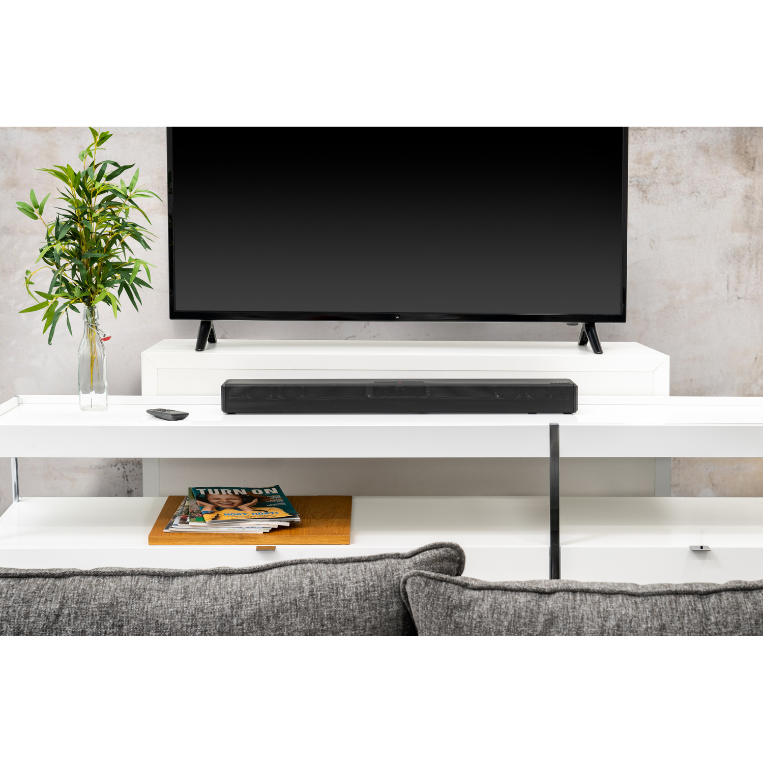 Een tv op een witte standaard met tijdschriften, een soundbar en een bank.