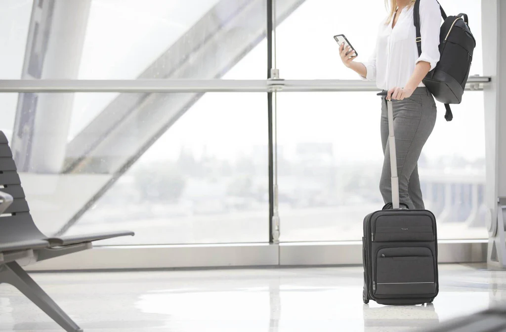 Mujer con ropa clara en aeropuerto, con equipaje y smartphone.