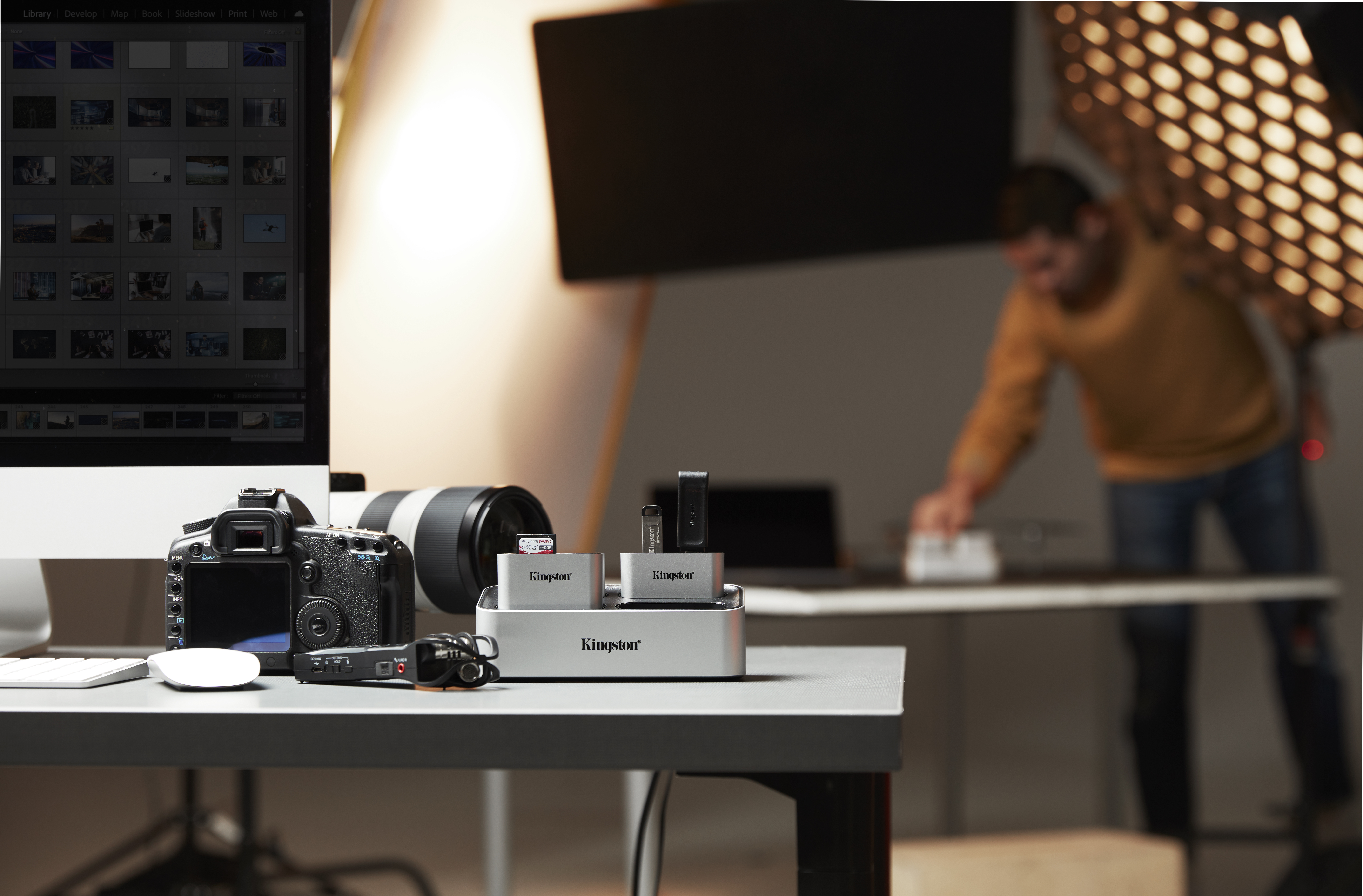 Ein Fotostudio mit Kamera, Objektiv und Zubehör auf dem Tisch; eine Person im Hintergrund.
