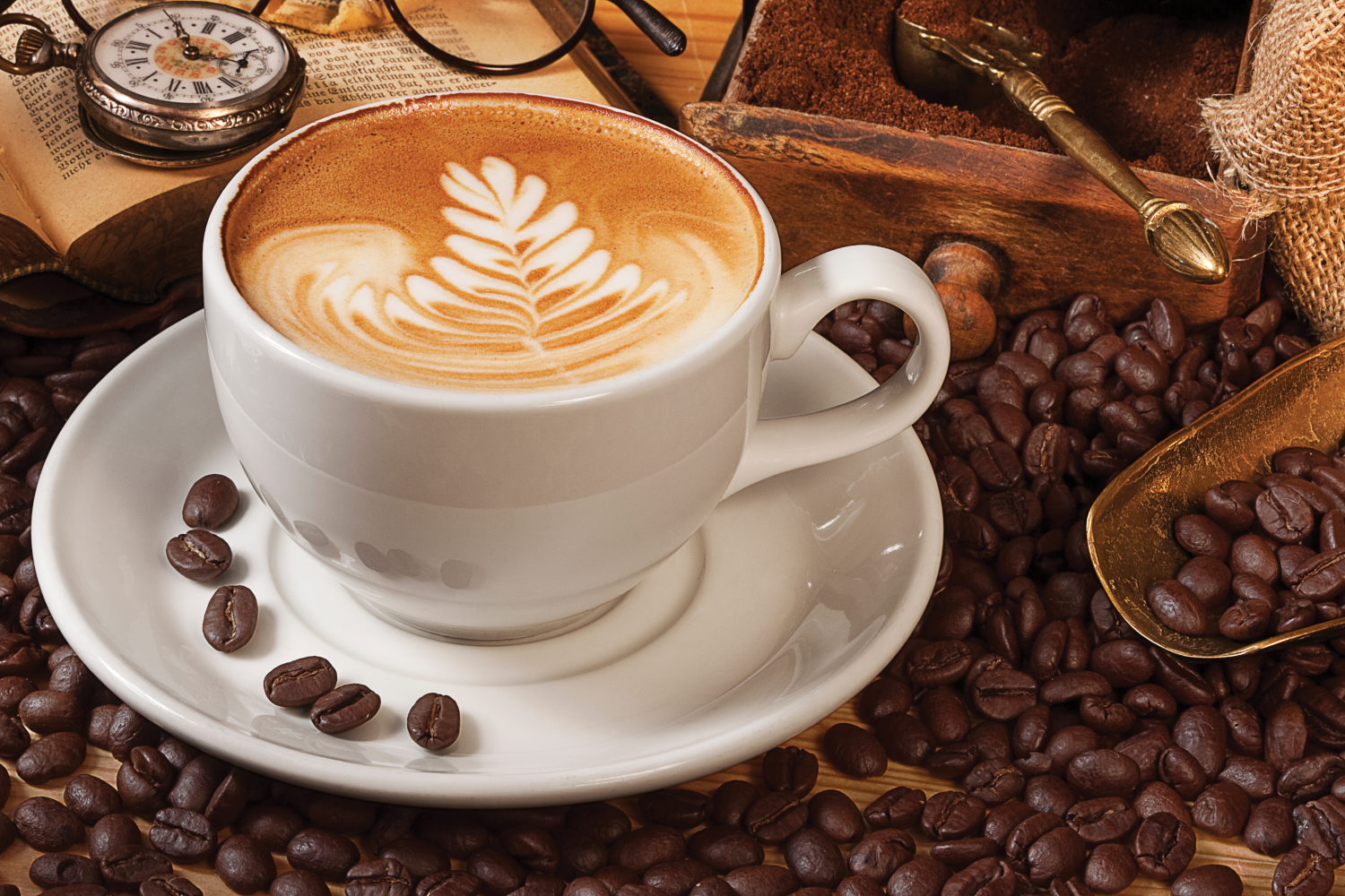 Latte egy fehér csészében csészealjjal, kávébabokkal körülvéve. Zsebóra és könyv az oldalon.