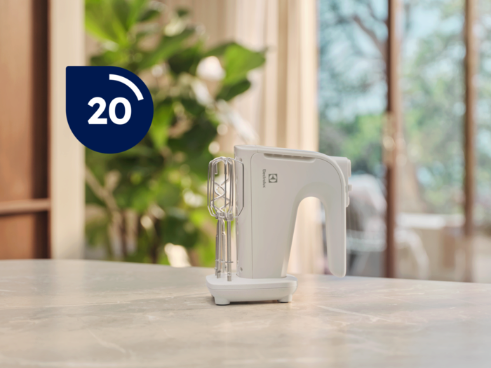 Fehér kézi mixer habverőkkel márványlapon. Kék kör 20-as számmal. Háttér: levelek és ablak.