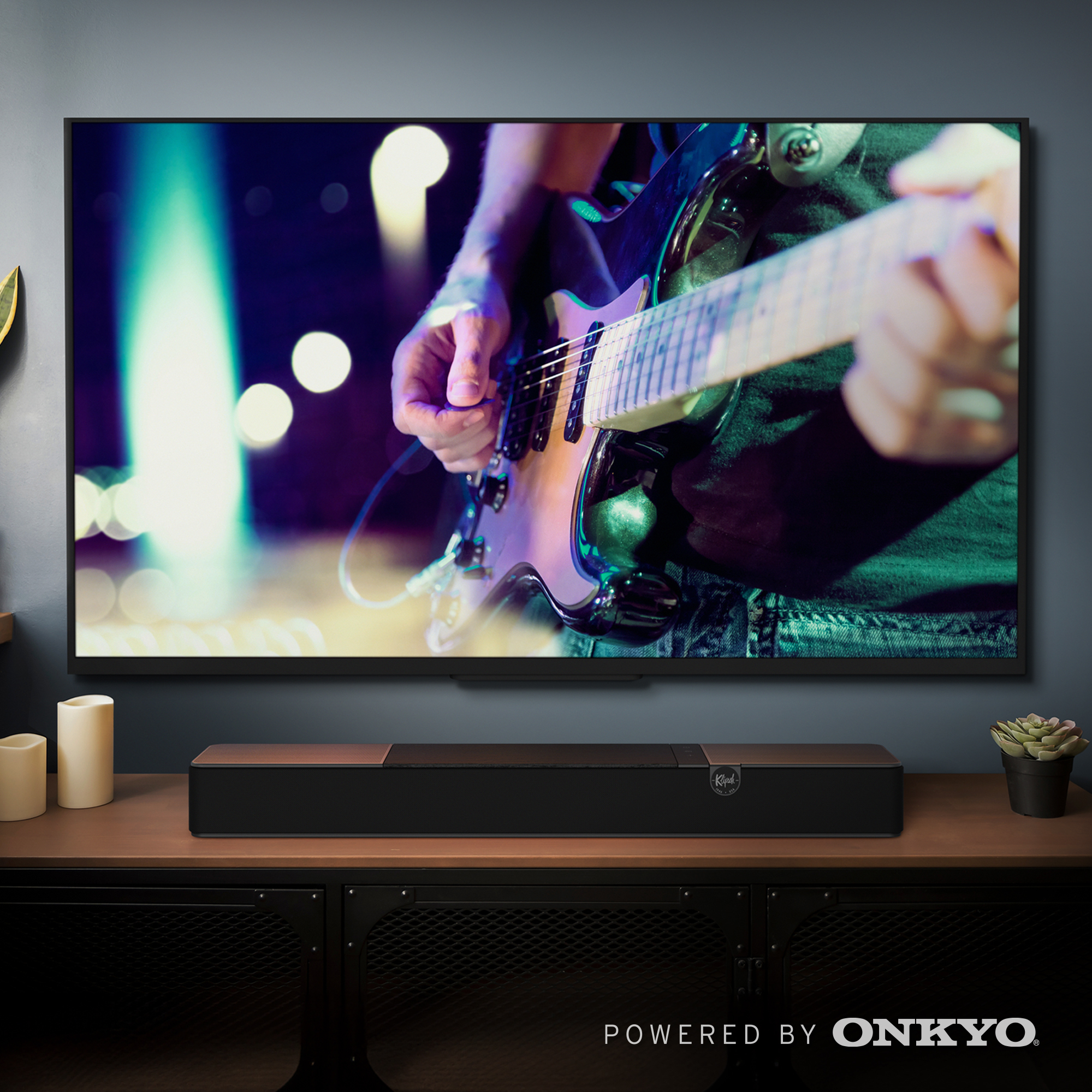 Ein Fernseher mit einem Gitarristen, darunter eine Soundbar auf einem Schrank, flankiert von Kerzen und einer Pflanze.