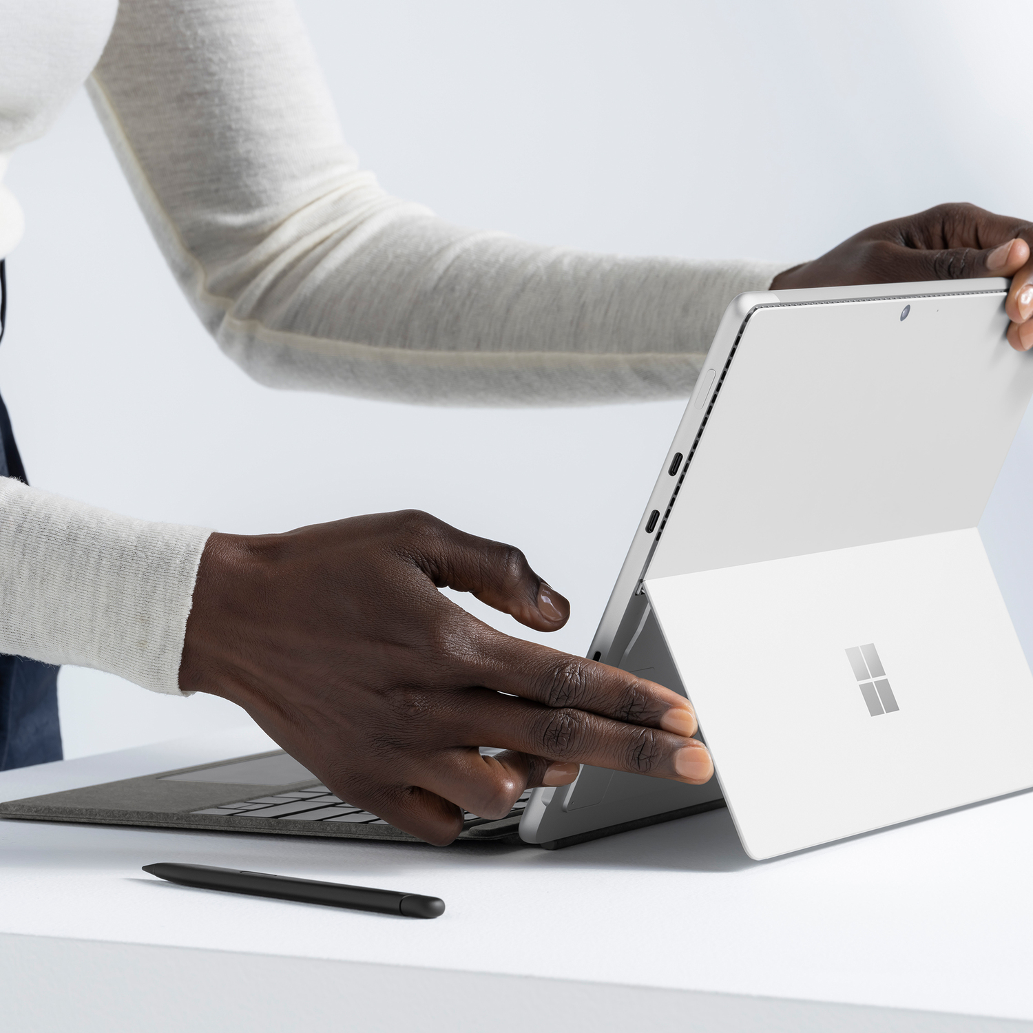 Handen die interageren met een zilveren laptop. De laptop is open. Een zwarte styluspen ligt op de tafel.