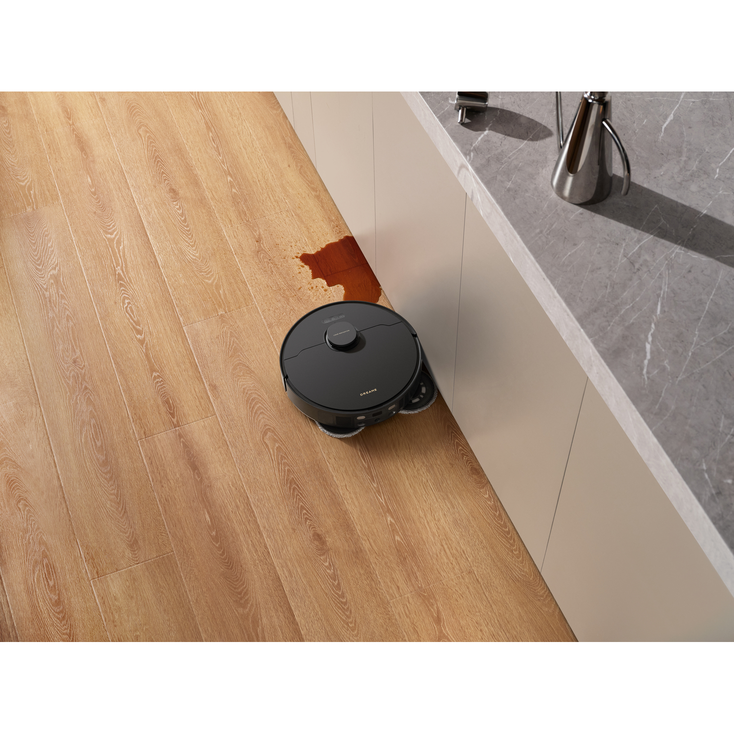 A black robot vacuum cleaner on wooden floor next to a cabinet. A brown stain is on the floor.