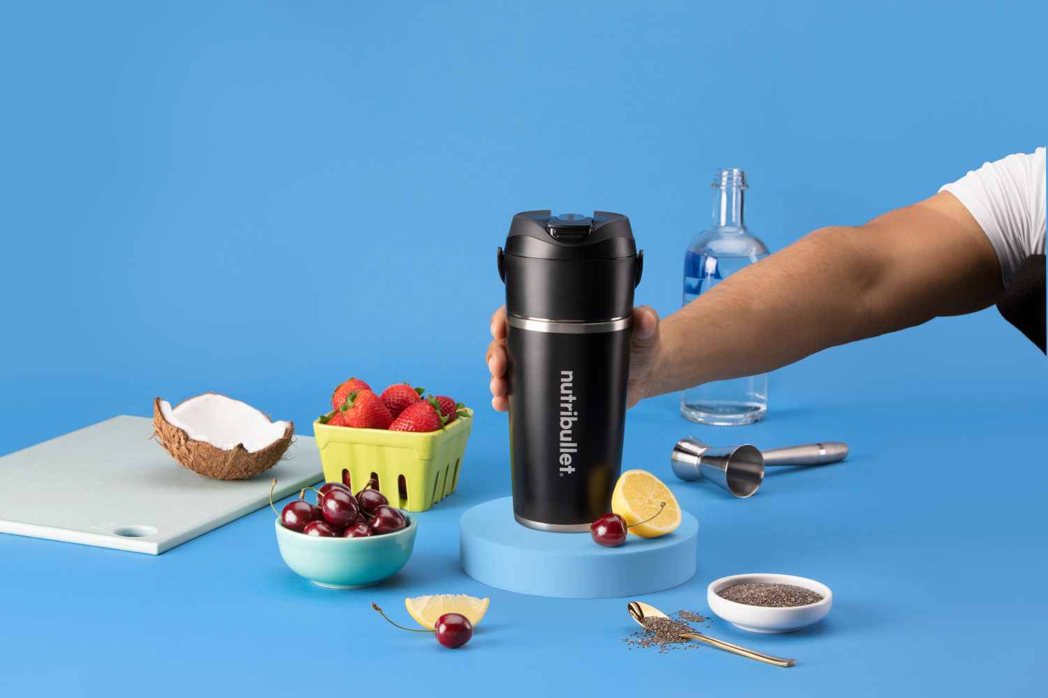Egy fekete Nutribullet csésze, amelyet egy kéz tart, gyümölcsökkel és italösszetevőkkel kék háttéren.