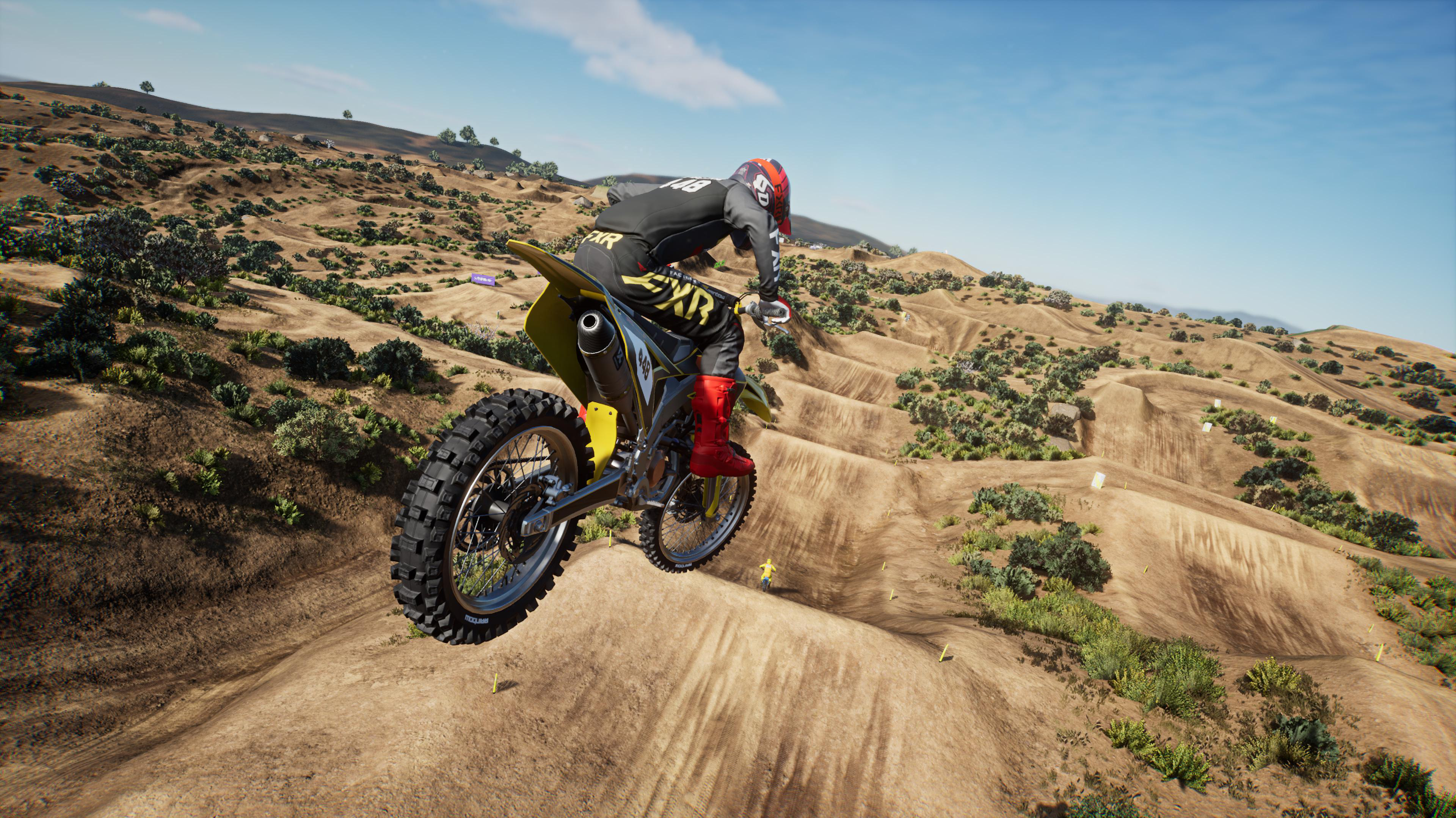 Ein Motocross-Fahrer auf einem gelben Motorrad springt über eine Dirt-Strecke, blauer Himmel im Hintergrund.