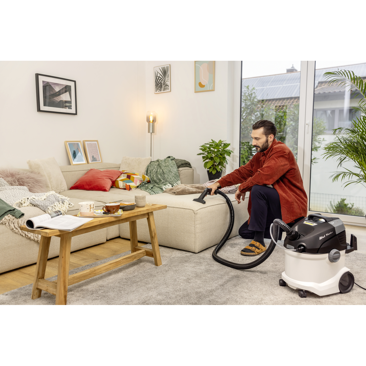 Man vacuuming a carpet in a living room with a sofa and windows.