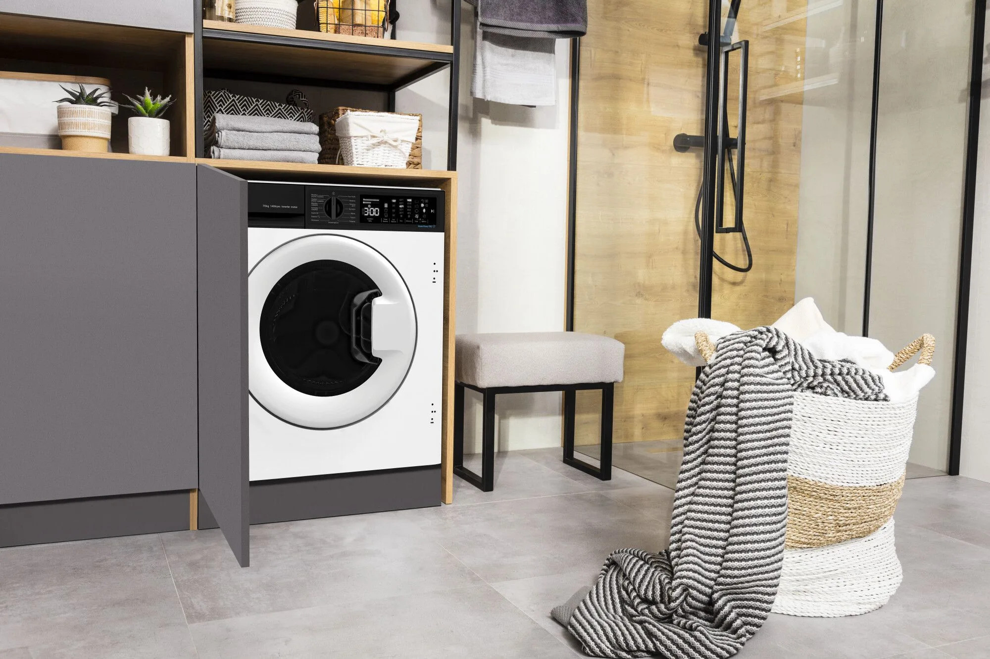 Biała pralka w łazience. Szara szafka, półki i stołek. Kosz z ręcznikami i dywanem.