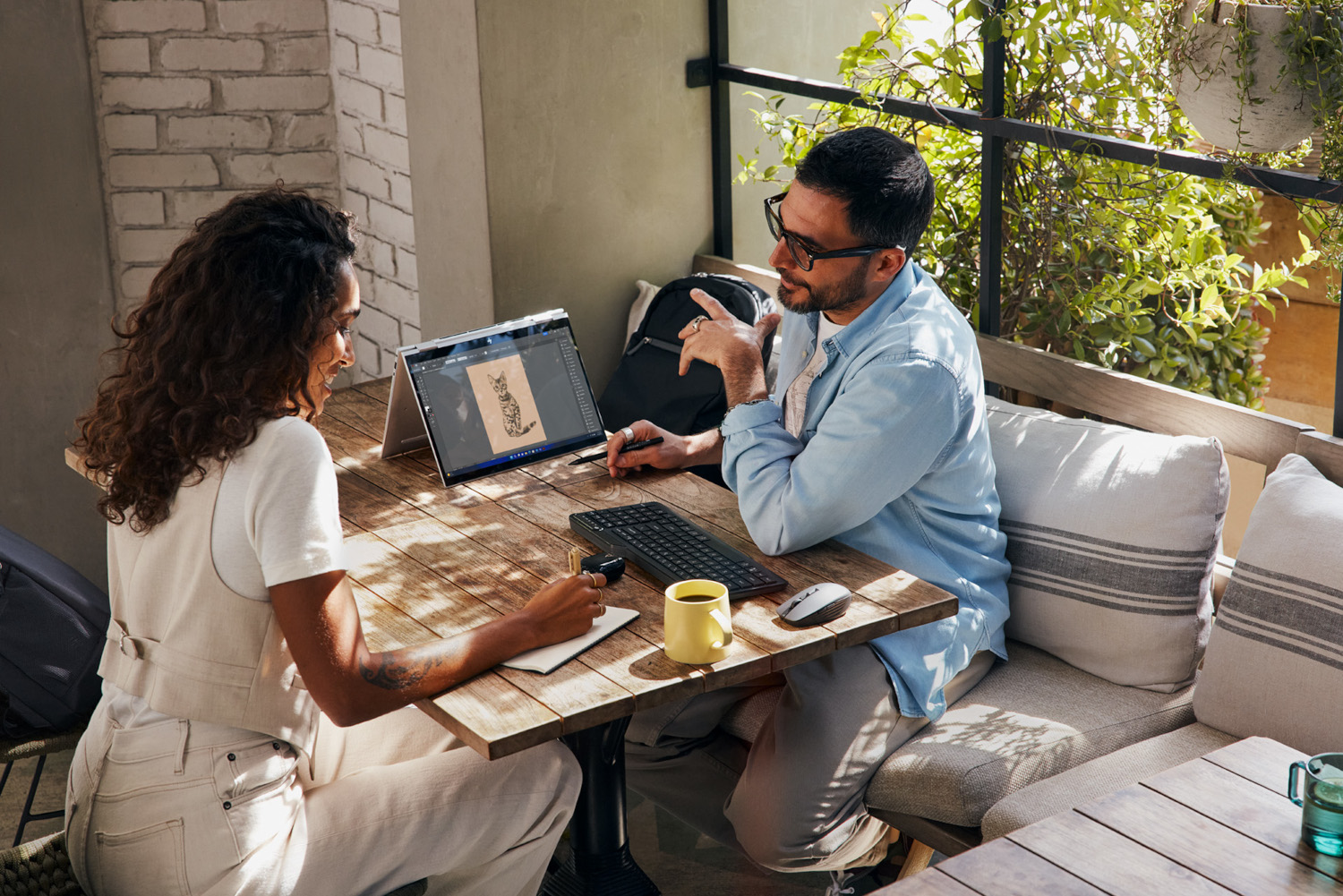 Two people at a wooden table with a laptop. A person writes on a notepad, and plants in the background.