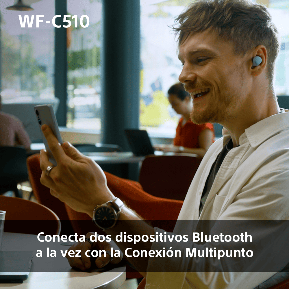 Un hombre sonríe mientras usa auriculares y sostiene un teléfono en un café.