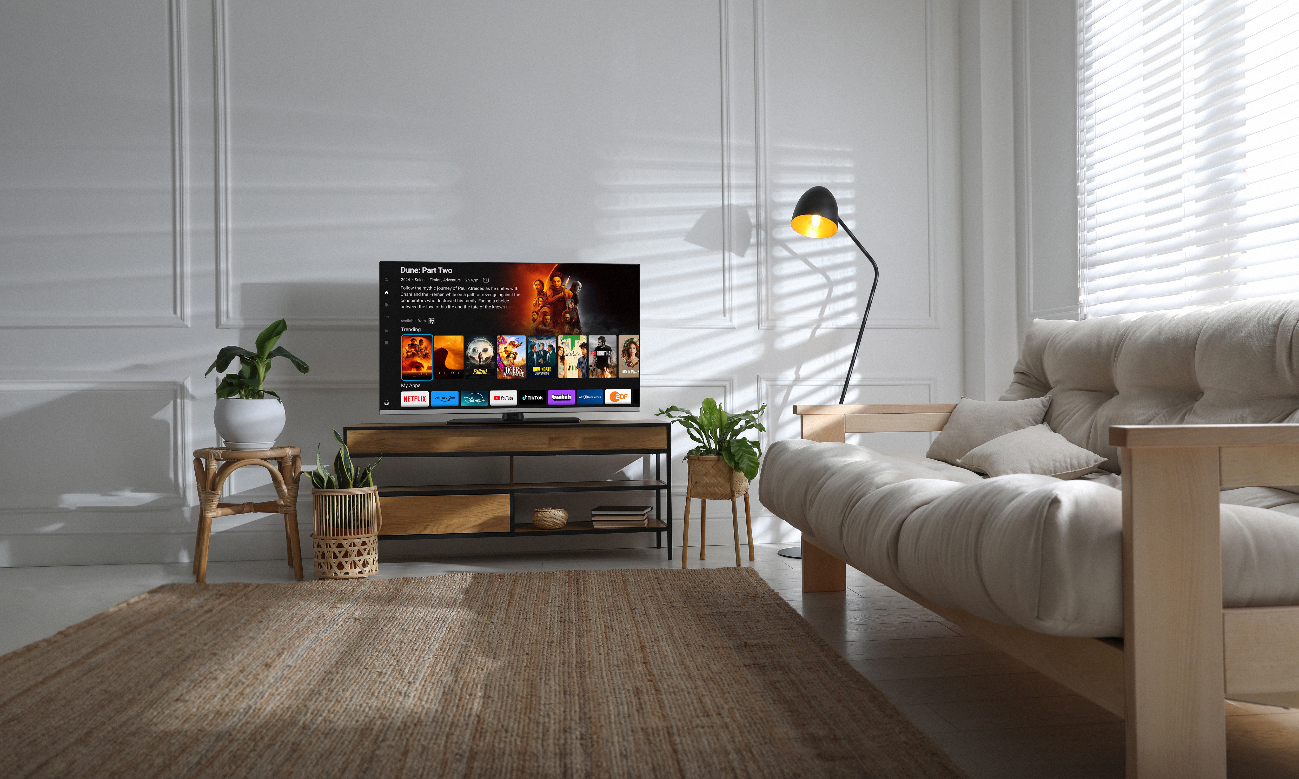 Salon avec télévision, canapé, tapis, plantes et lampadaire, créant une atmosphère chaleureuse et bien éclairée.
