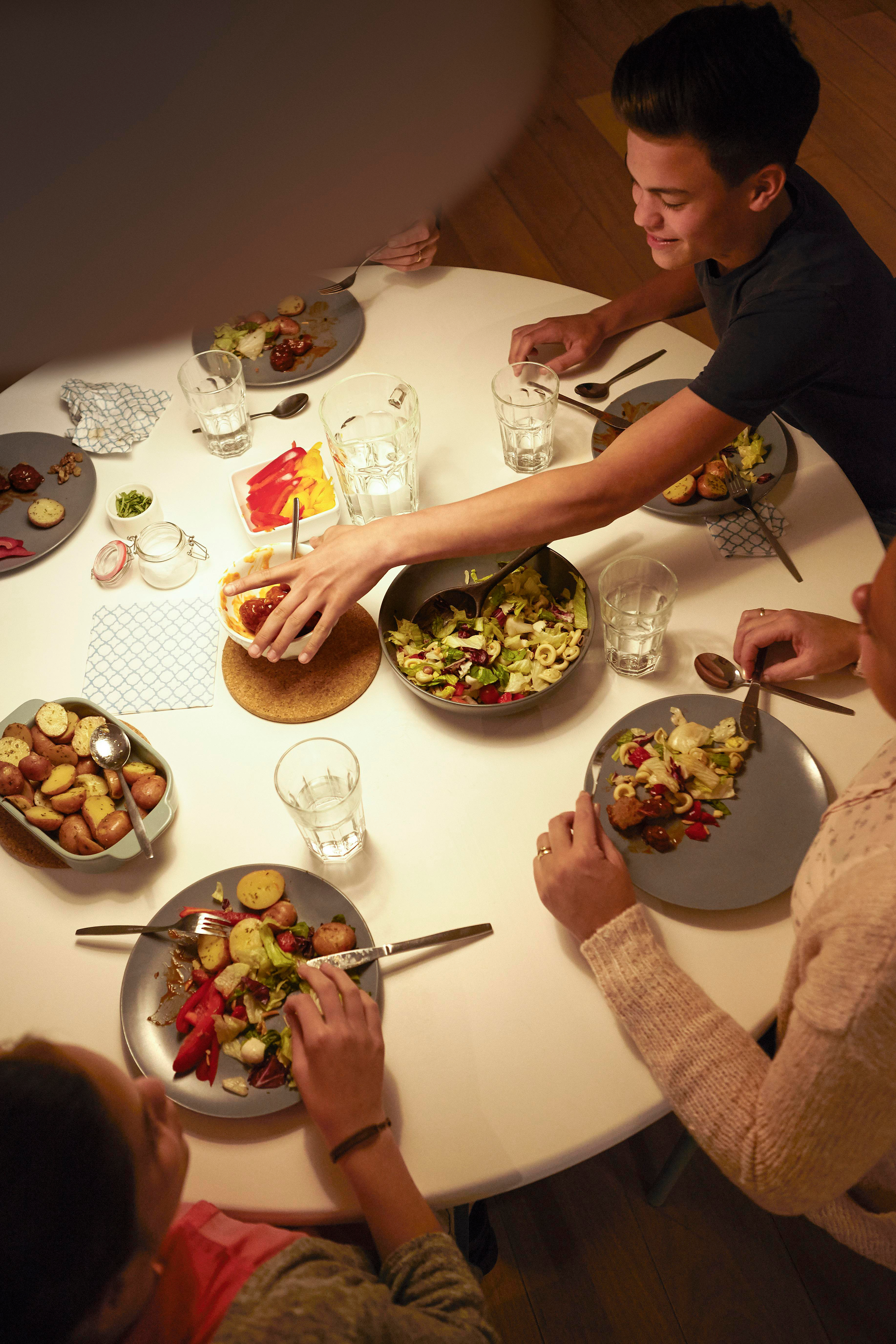 Menschen an einem weißen Tisch mit Essen auf Tellern, Hände greifen danach.