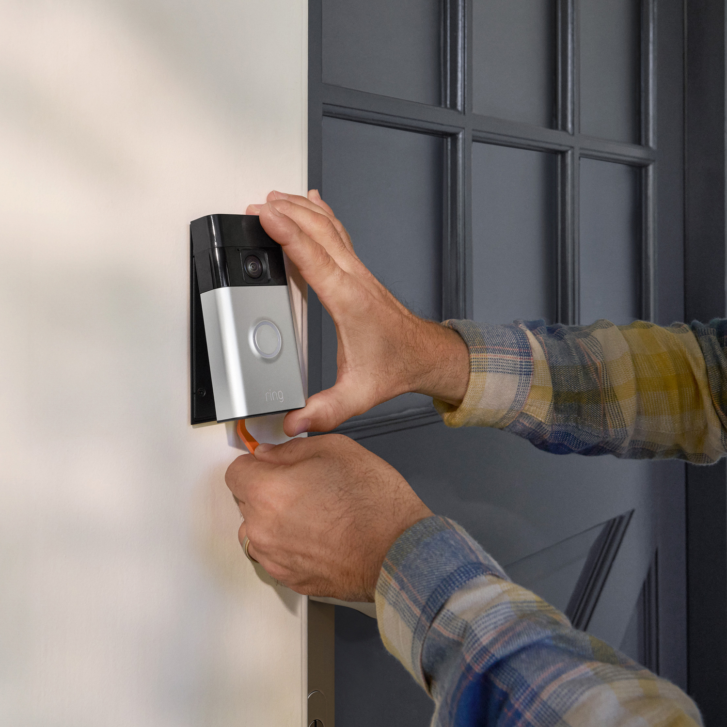 A person installing a silver smart doorbell on a white wall next to a dark gray door.