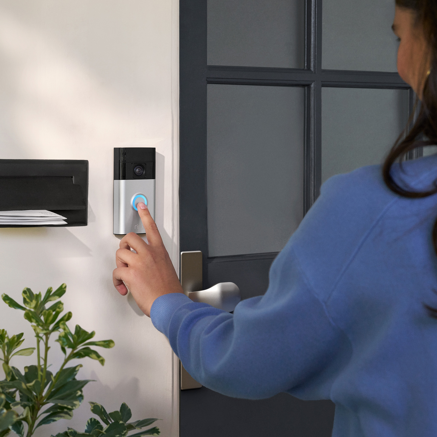Woman pressing a doorbell button on a wall. Door and a mailbox are nearby.