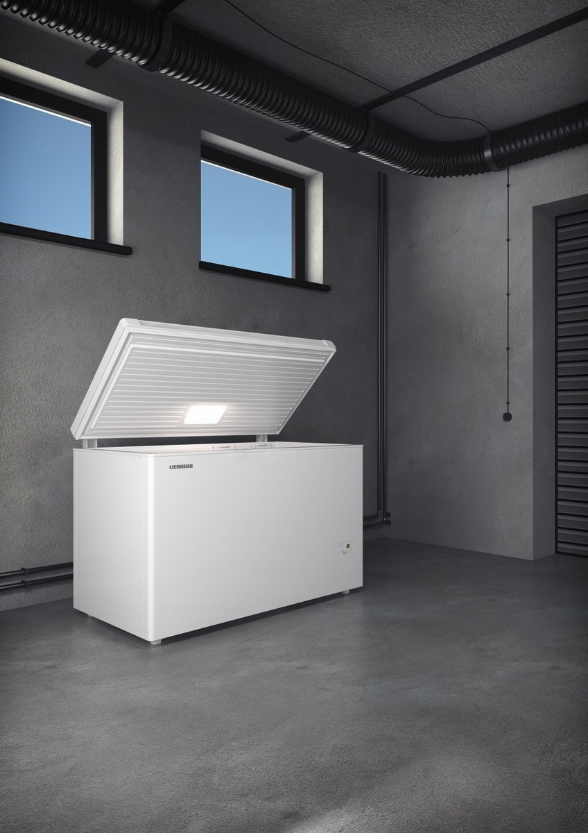 Biała zamrażarka z otwartą pokrywą w szarym pomieszczeniu z dwoma oknami i czarną rurą.