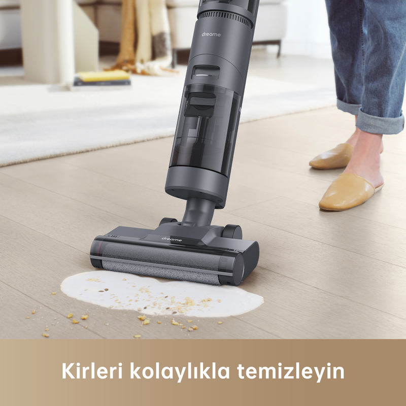 Gri bir elektrikli süpürge, ahşap zemindeki bir döküntüyü temizliyor. Arka planda bir kişi var.
