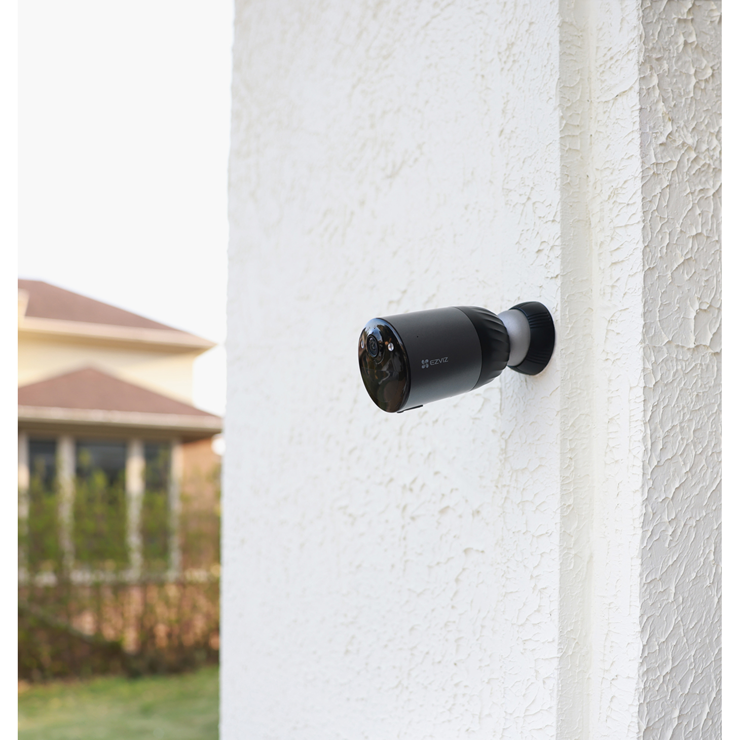 Black security camera mounted on a white textured wall outside, with a house visible.