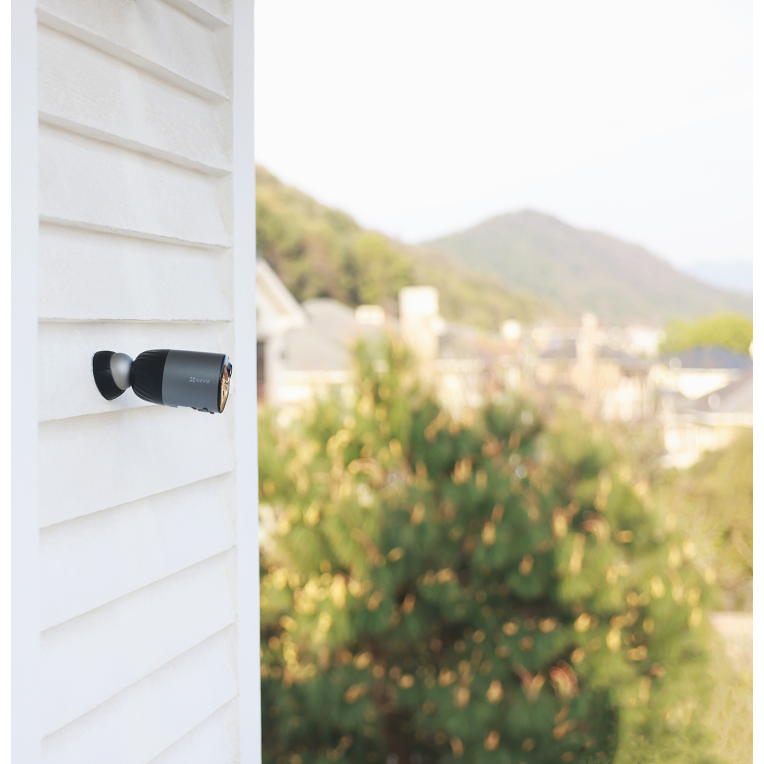 Outdoor security camera mounted on a wall. Background: trees, houses, and mountains.