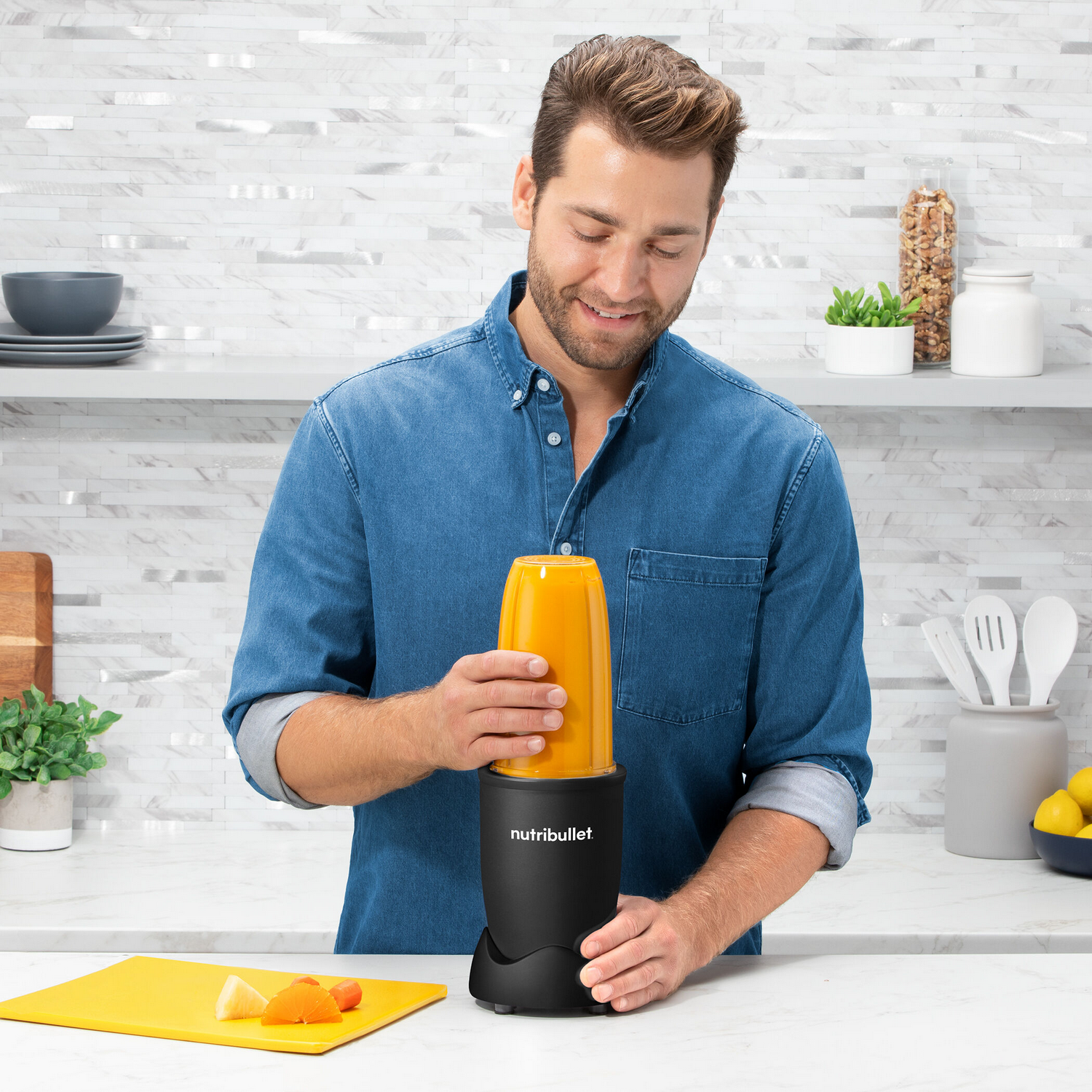 Hombre usando una licuadora Nutribullet negra, sosteniendo un recipiente amarillo con contenido mezclado.