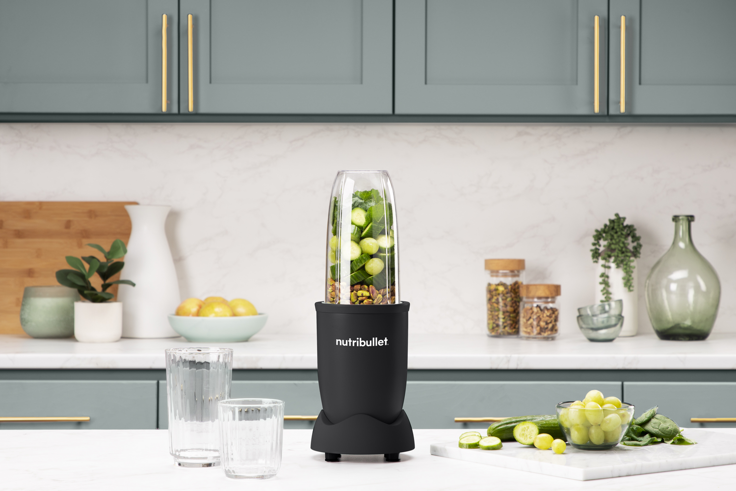 Licuadora negra con ingredientes y vasos en una superficie blanca.