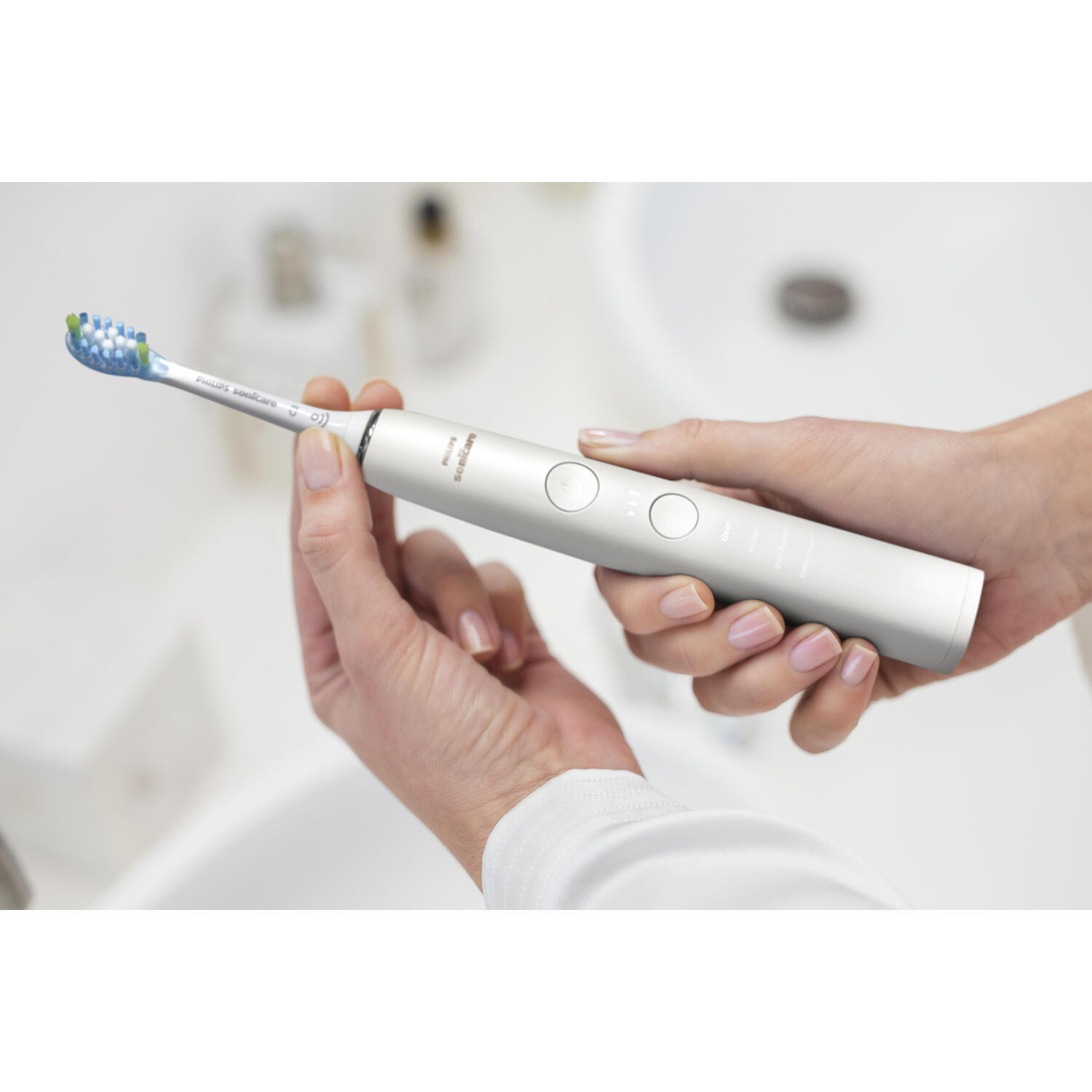Une personne tient une brosse à dents électrique blanche et argentée. Évier en arrière-plan.