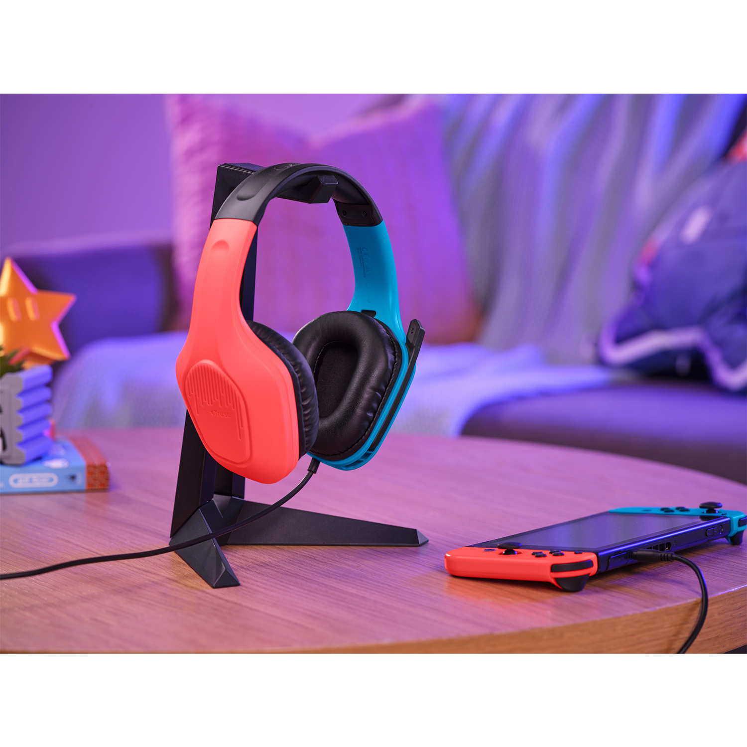 Cuffie da gioco rosse e blu su un supporto con Switch e controller. Sfondo sfocato.