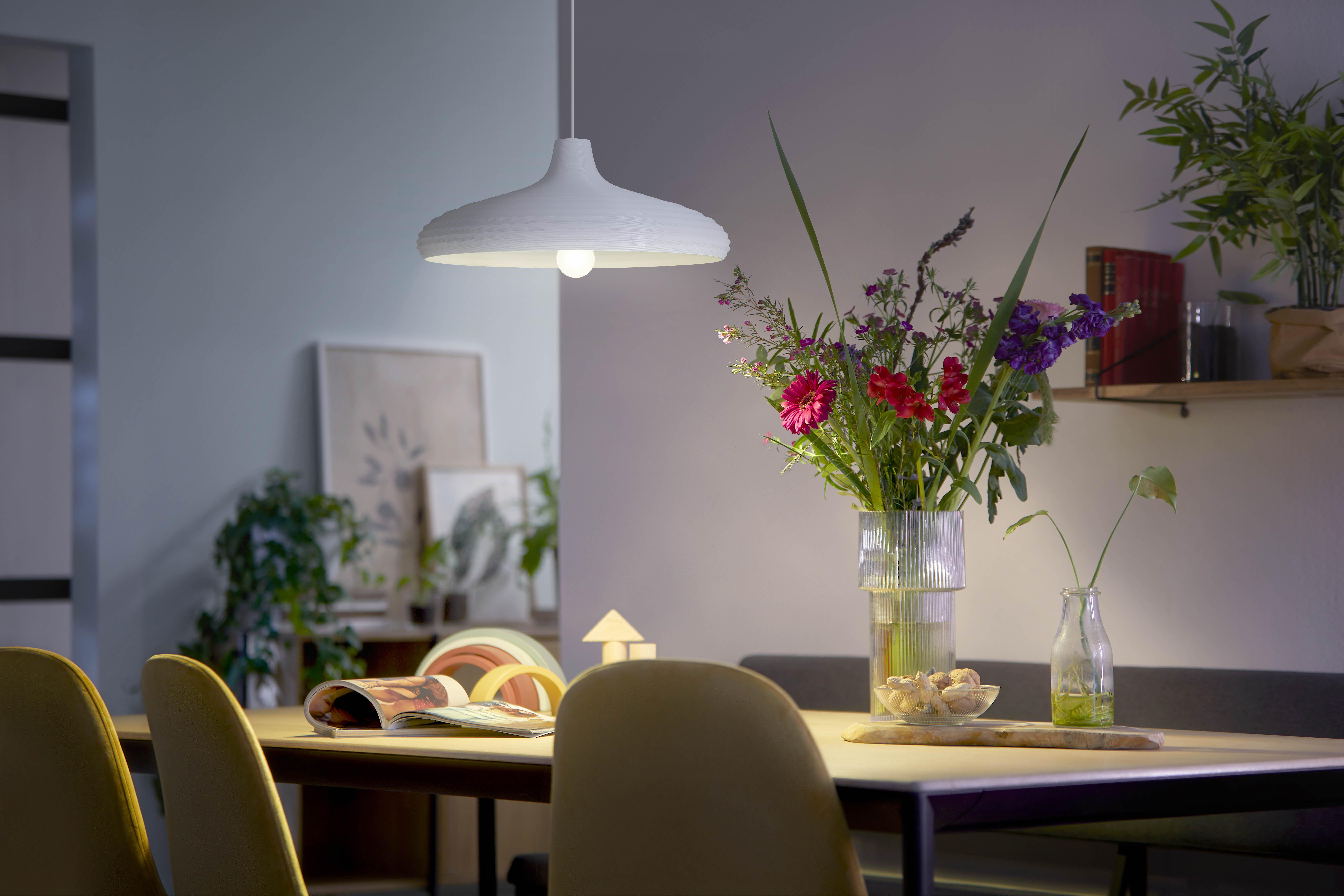 Ein Esstisch mit Blumen, Büchern und einer Lampe. Der Raum ist beleuchtet.