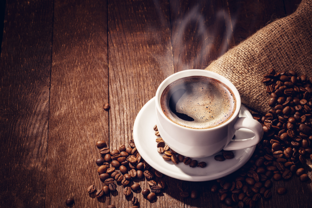 Steaming cup of coffee on a saucer, surrounded by scattered coffee beans on a wooden surface.