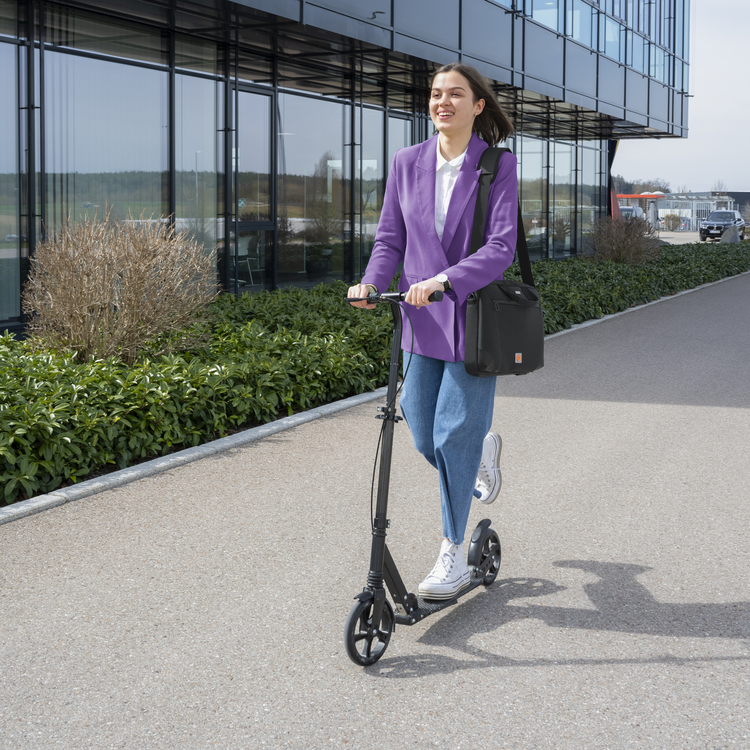 Vrouw op een zwarte scooter op een stoep. Ze draagt een paarse blazer en jeans. Een zwarte tas hangt aan haar schouder.