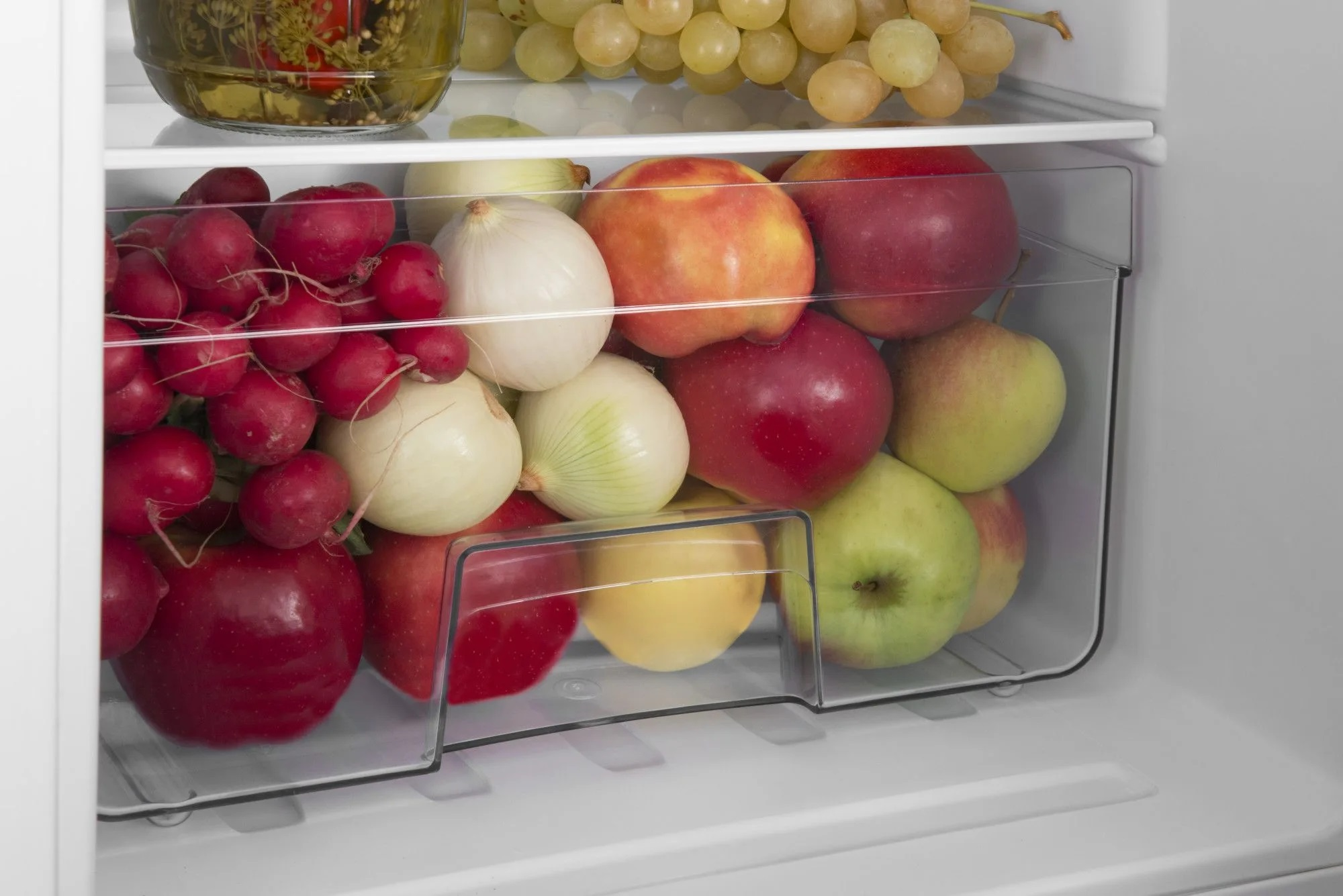 Szuflada lodówki wypełniona owocami i warzywami: rzodkiewki, cebula, jabłka i winogrona.