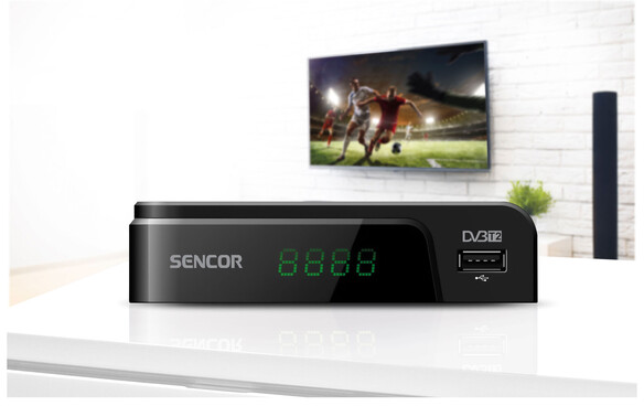 Fekete Sencor DVB-T2 vevő zöld kijelzővel. TV a háttérben.
