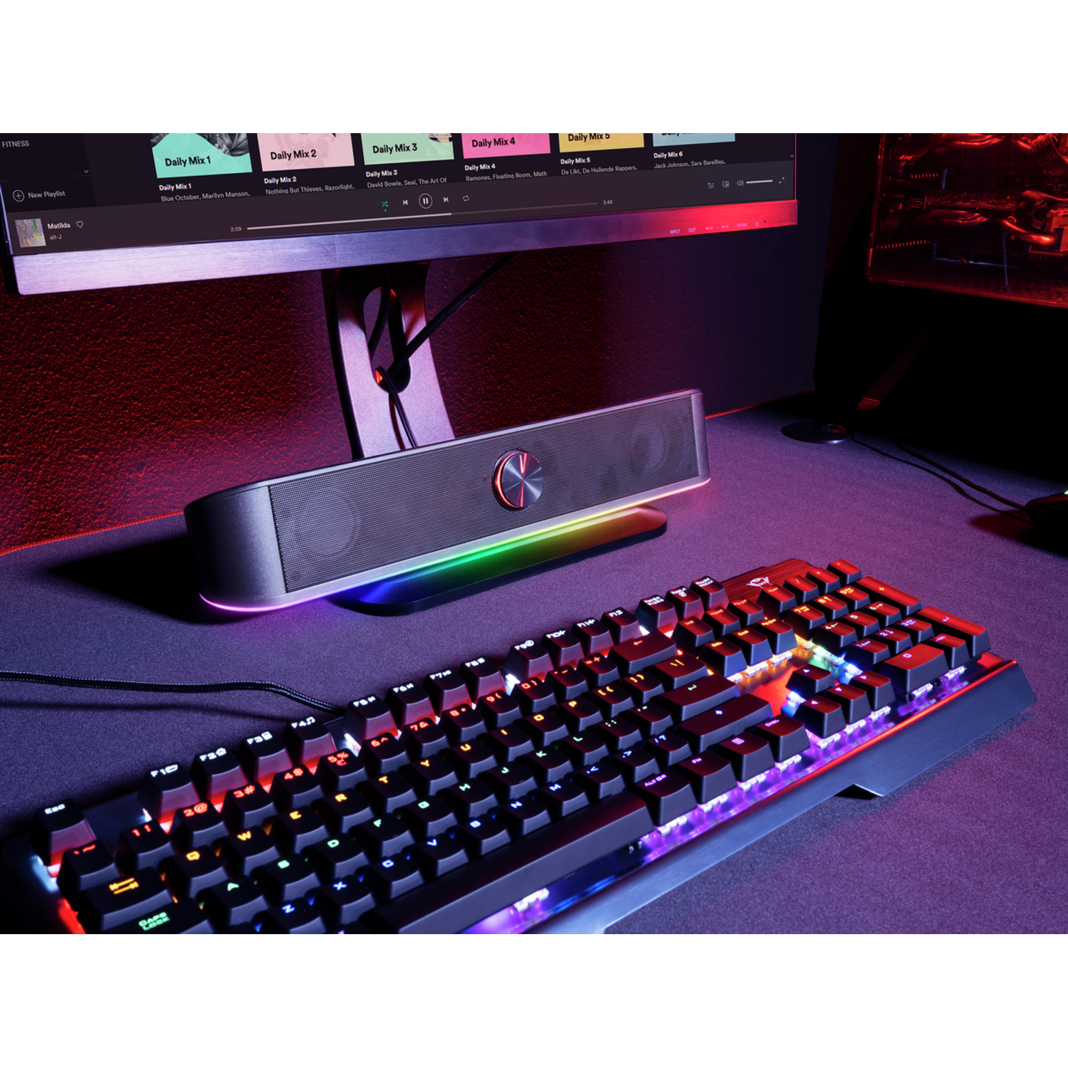 Lit keyboard, speaker, and monitor. Keyboard keys glow. Speaker has a rainbow light. Monitor shows a music playlist.