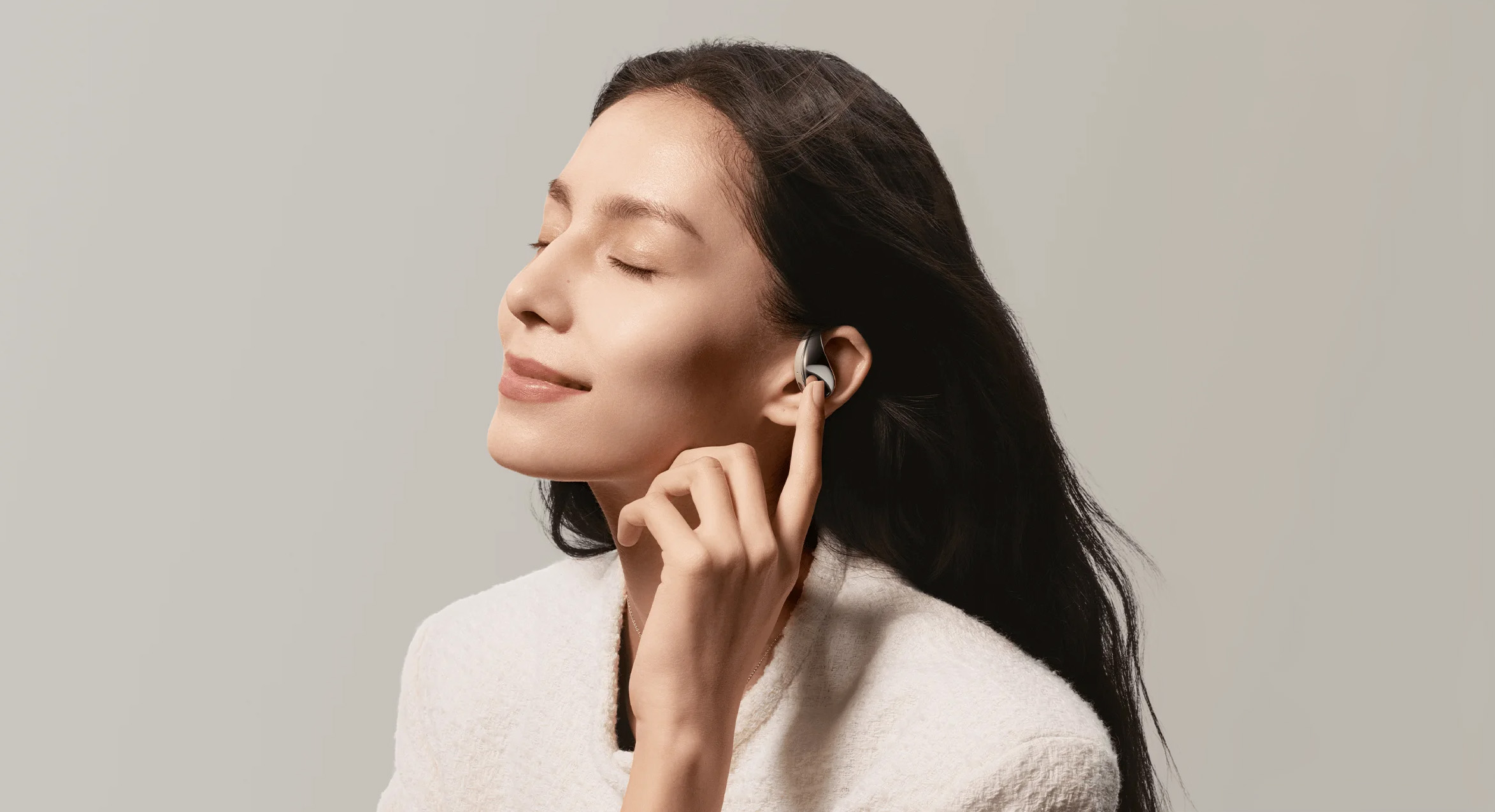 Kobieta nosząca słuchawki w uszach. Dotyka jednej słuchawki. Ma długie ciemne włosy i białą kurtkę.