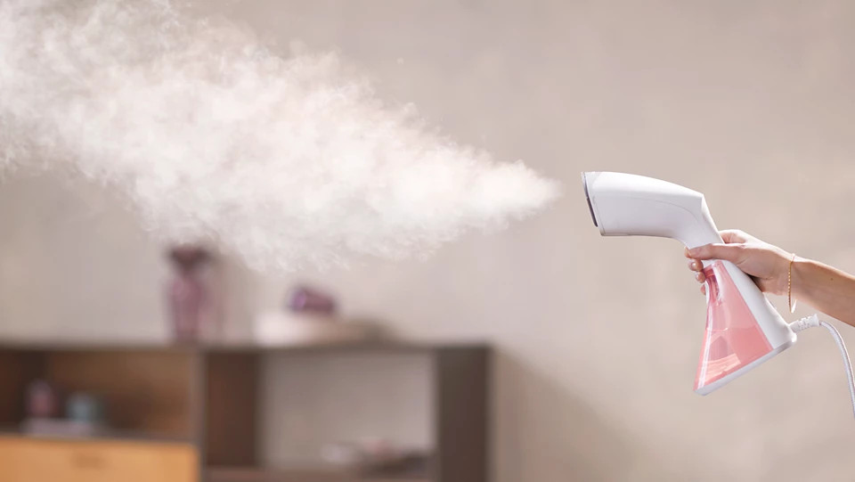 Dłoń trzymająca biało-różowy parownik do ubrań, wydobywająca parę.