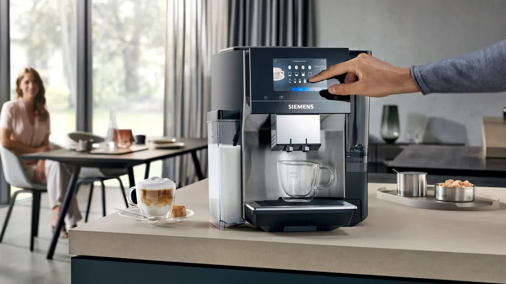 Egy személy kávéfőző érintőképernyőjét használja. Egy csésze és egy latte a pulton.