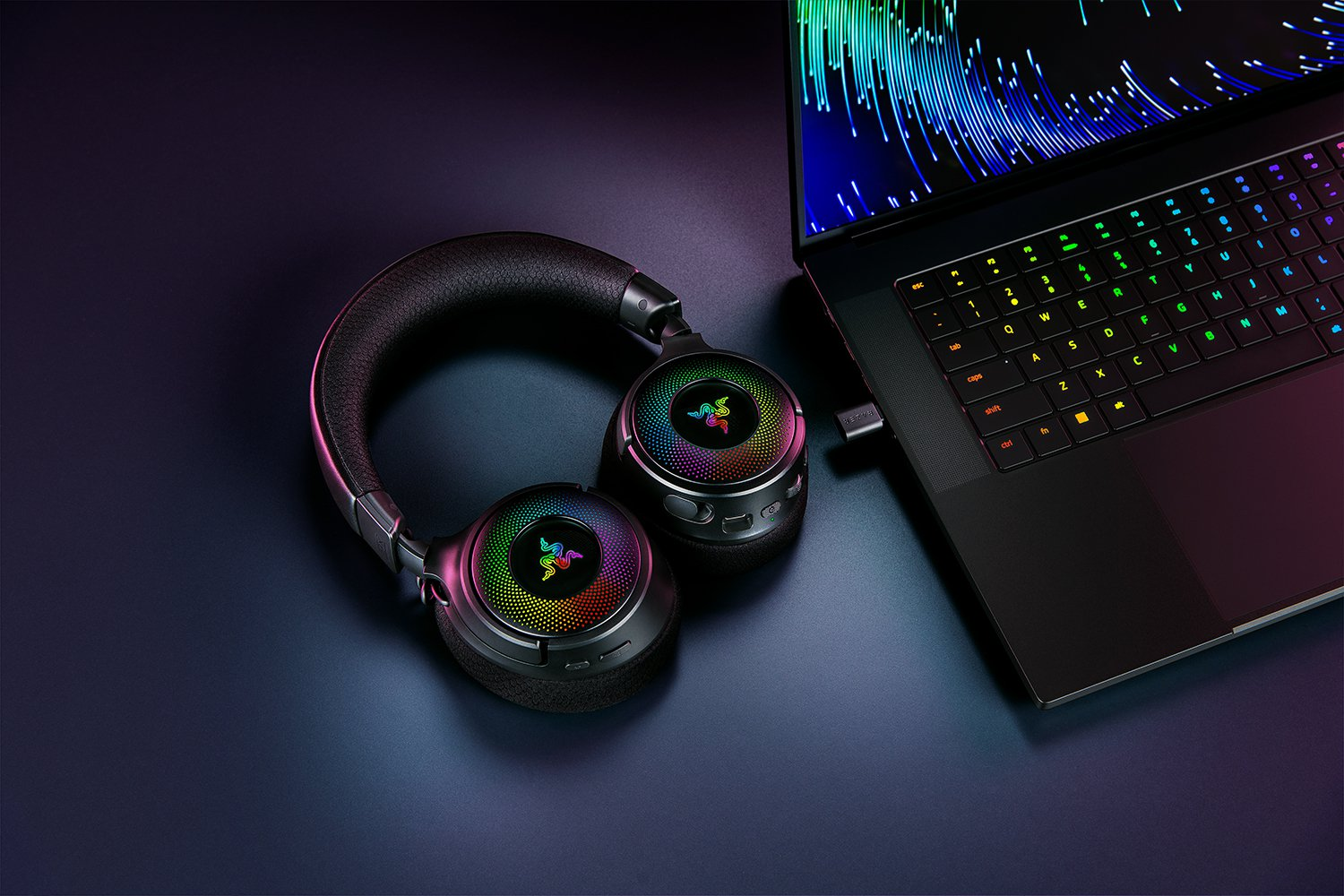 Cuffie Razer nere e un adattatore USB accanto a un laptop che mostra una grafica astratta e colorata.