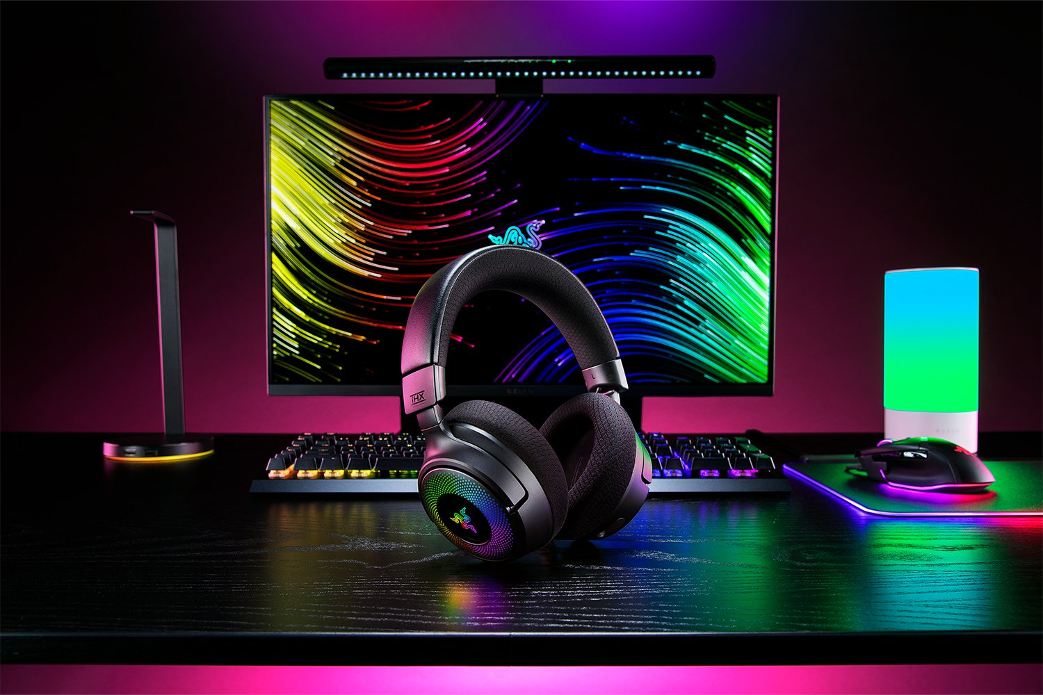 Configurazione gaming: cuffie, monitor, tastiera, mouse e lampada da scrivania su una scrivania scura, con illuminazione colorata.