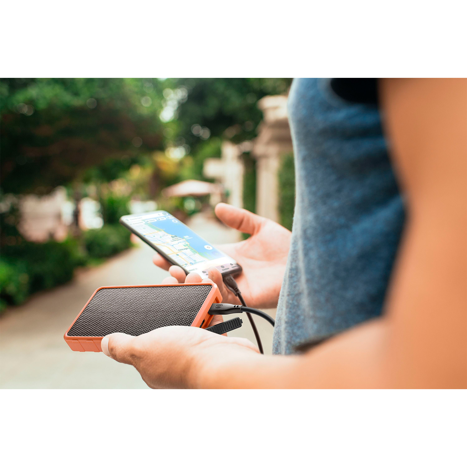 Persona che tiene in mano un altoparlante arancione e uno smartphone con mappa. Ambiente esterno, sfondo sfocato.