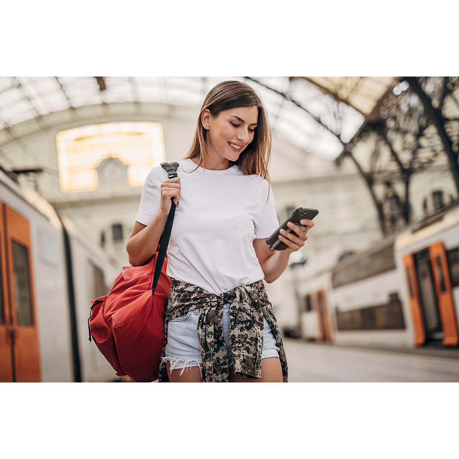 Donna in stazione che guarda il telefono, con borsa rossa e treno in vista.