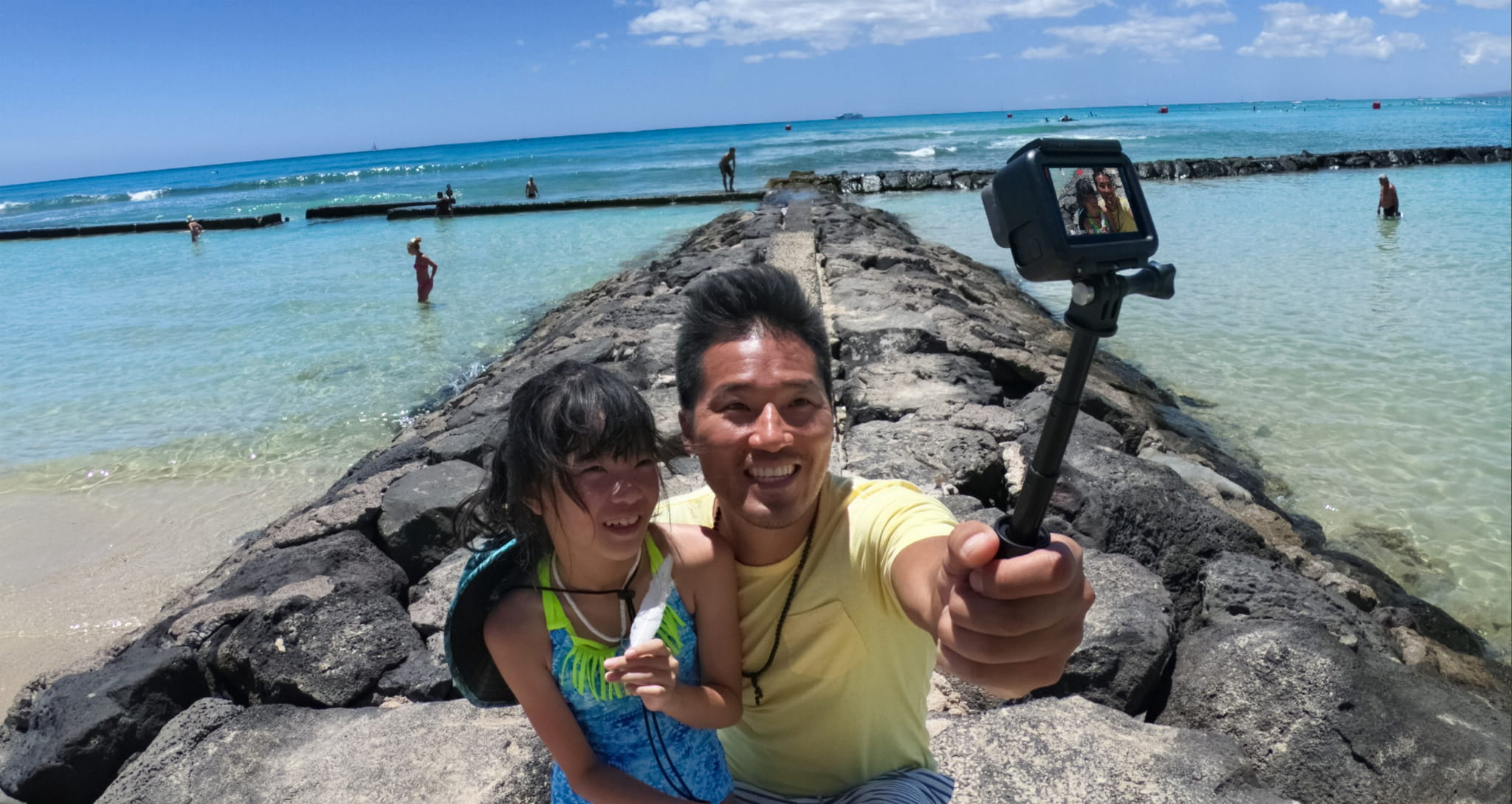 Mężczyzna i dziewczynka robią sobie selfie na kamiennej ścieżce na plaży. Czysta woda, błękitne niebo i inni ludzie w tle.