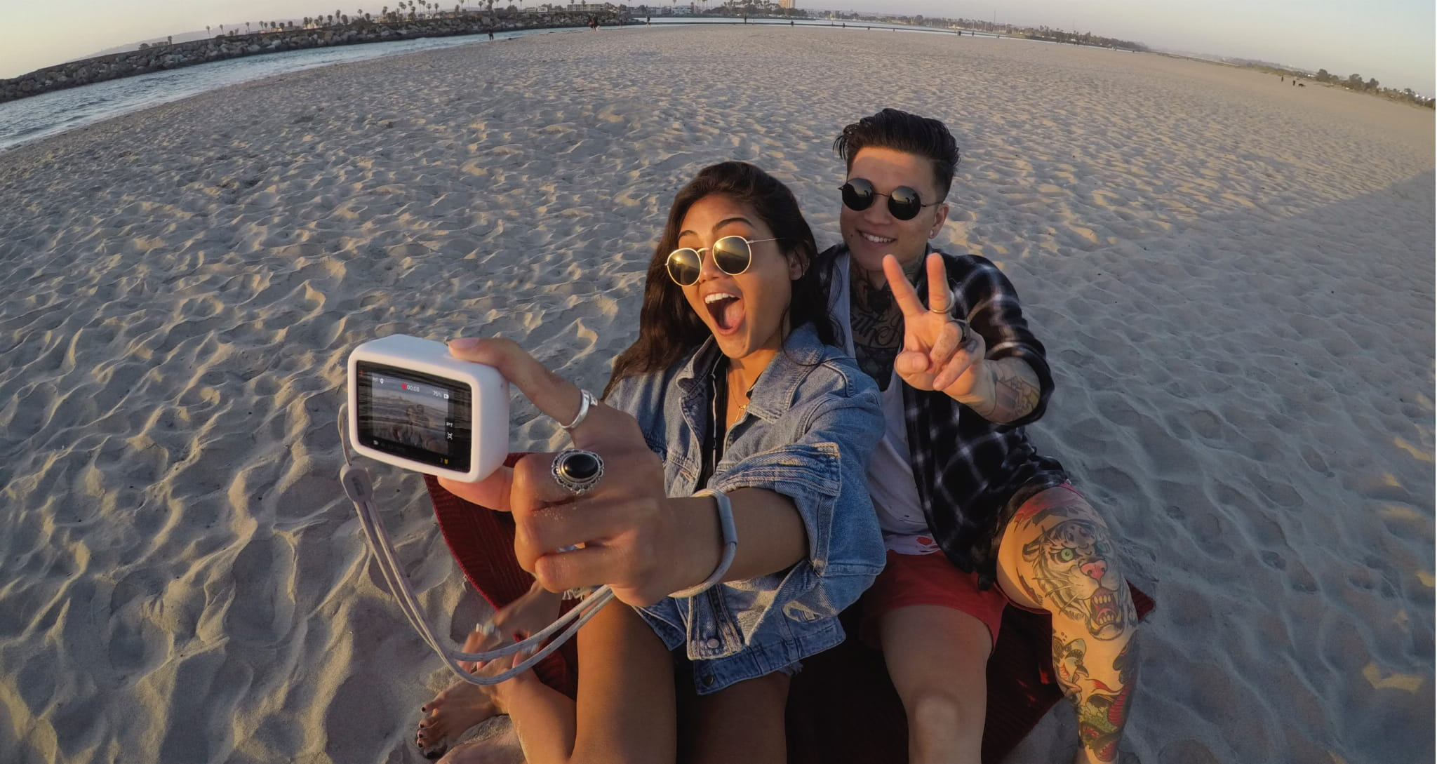 Dwie osoby robią sobie selfie na plaży. Jedna trzyma aparat, uśmiechając się. Druga pokazuje znak pokoju, z tatuażami. Tło piaszczystej plaży.