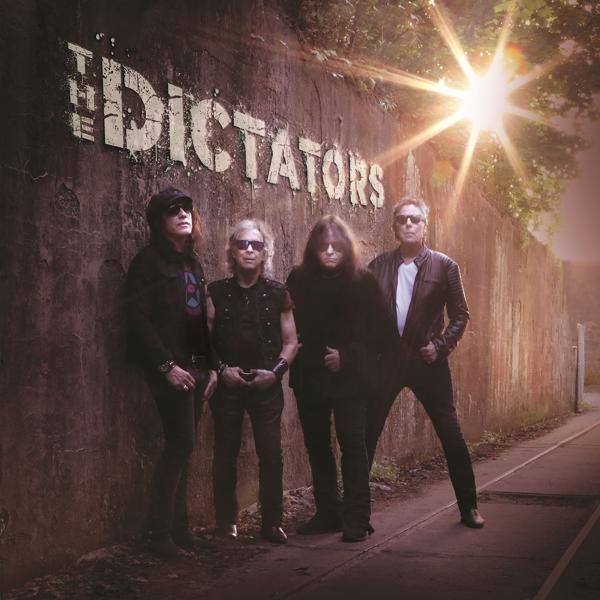 Le groupe The Dictators pose devant un mur de briques. Le soleil brille en arrière-plan.