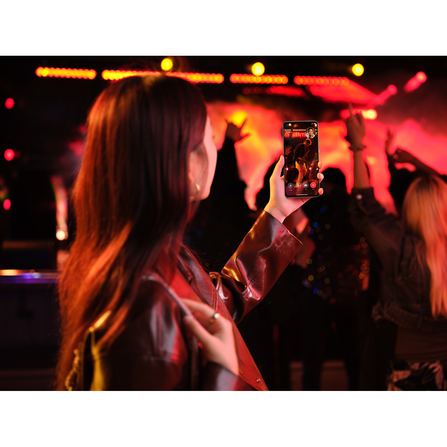 A woman holding a phone at a concert. The screen shows a person on stage.