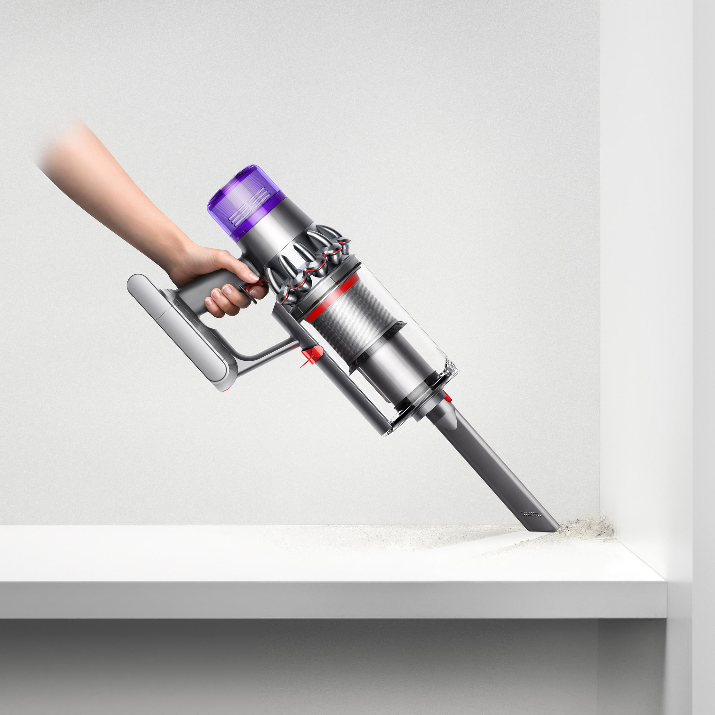 Eine Hand, die einen silbernen Dyson-Staubsauger auf einem weißen Regal benutzt, um Staub zu entfernen.